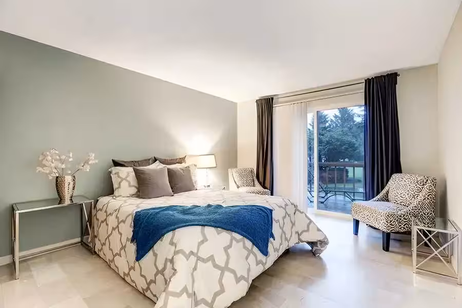 A master bedroom showcasing a pale green accent wall, queen size bed topped beddings, two armchairs next to the door